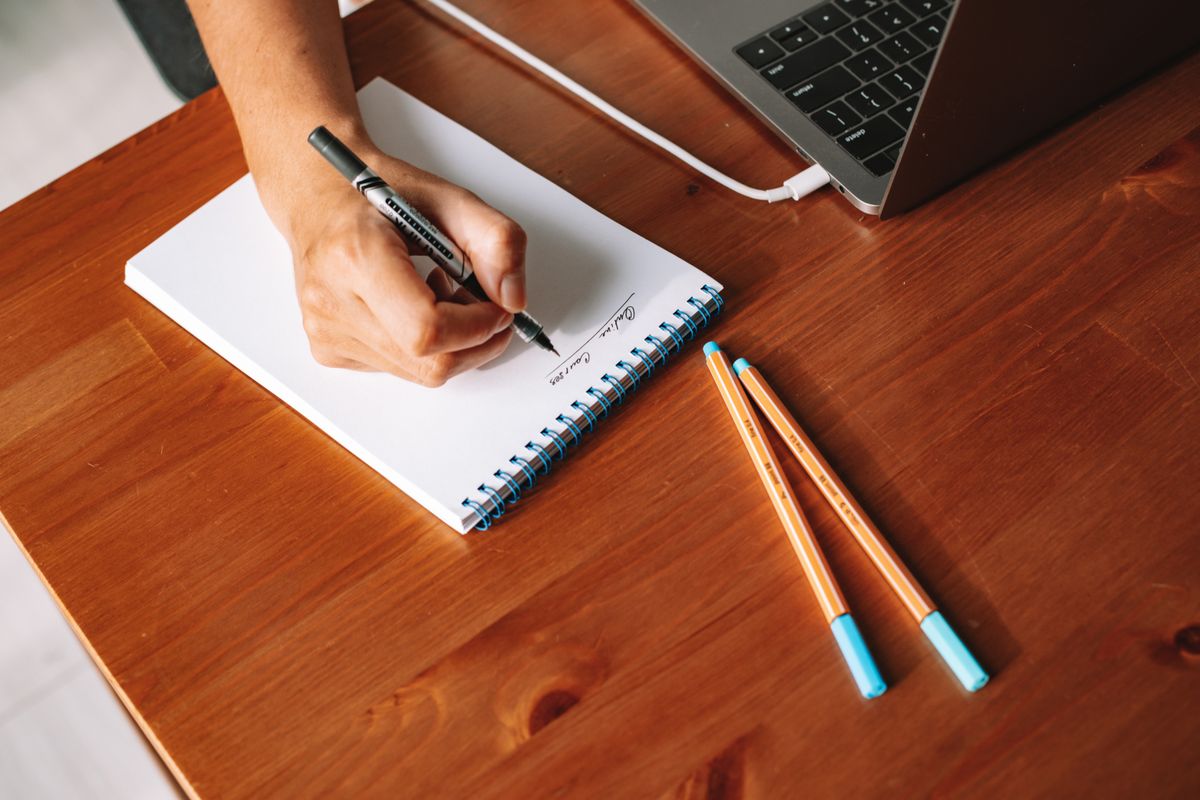 A hand writing in a notebook beside a laptop